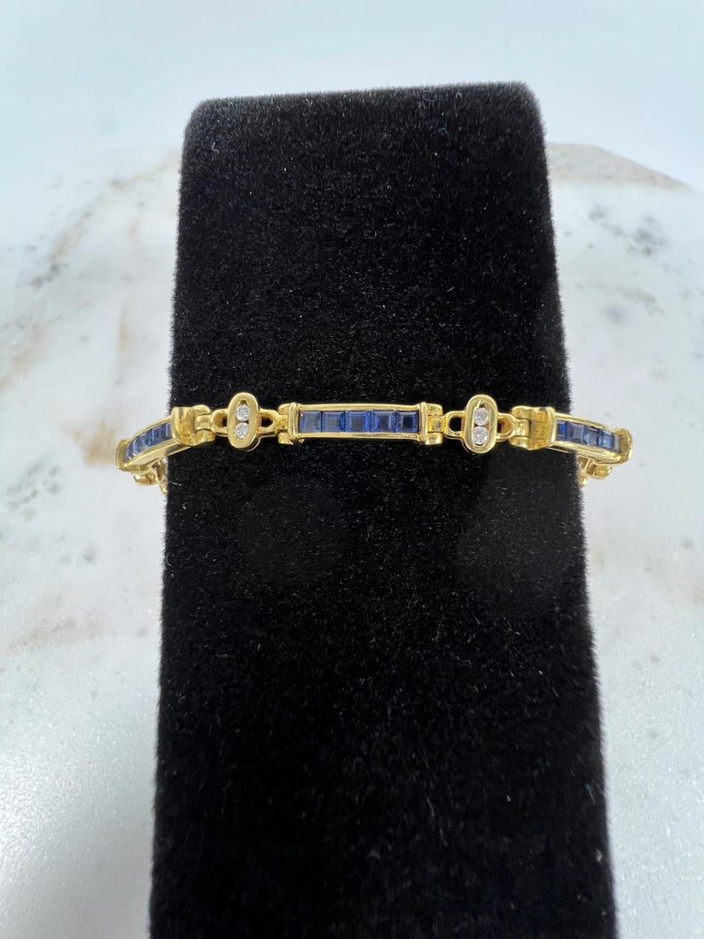 May include: A gold link bracelet featuring a series of square-cut blue gemstones set in a channel style, interspersed with small oval-shaped gold links that each contain two tiny round clear stones arranged vertically along the center.