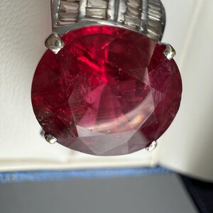 May include: A ring featuring a large, oval-cut, deep red gemstone held by four silver-toned prongs. The band is crafted from a silver-toned metal and is embellished with a row of small, rectangular, clear baguette-cut stones along the top section.