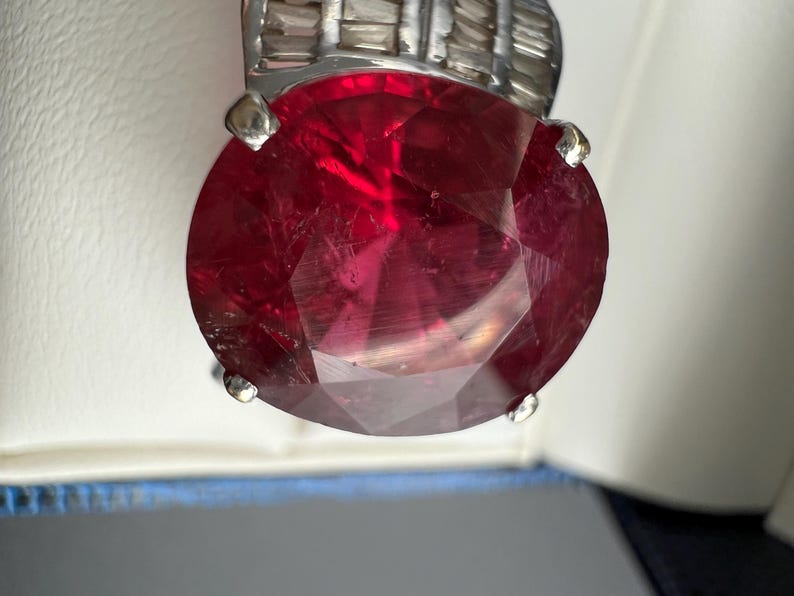 May include: A ring featuring a large, oval-cut, deep red gemstone held by four silver-toned prongs. The band is crafted from a silver-toned metal and is embellished with a row of small, rectangular, clear baguette-cut stones along the top section.