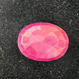 May include: A vibrant pink oval-shaped gemstone with a faceted cut. The gemstone is set against a black background.