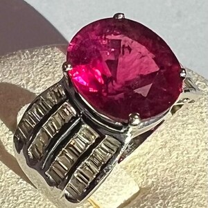 May include: A silver ring featuring a large, oval-cut, deep pink gemstone set in a four-prong mount. The band is wide and constructed with three parallel, horizontal rows of small, rectangular, clear stones embedded in the metal surface.