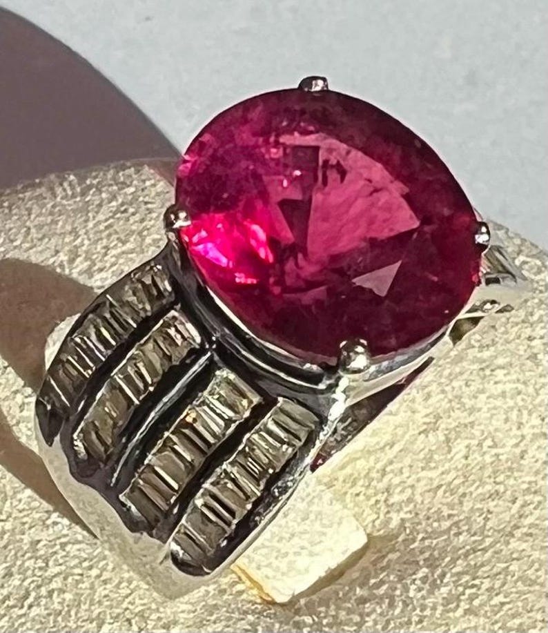 May include: A silver ring featuring a large, oval-cut, deep pink gemstone set in a four-prong mount. The band is wide and constructed with three parallel, horizontal rows of small, rectangular, clear stones embedded in the metal surface.