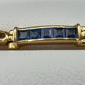 May include: A gold link bracelet featuring alternating sections of channel-set square blue sapphires and oval-shaped gold links, each containing two small round clear diamonds. The bracelet has a polished gold finish and a repeating geometric design.