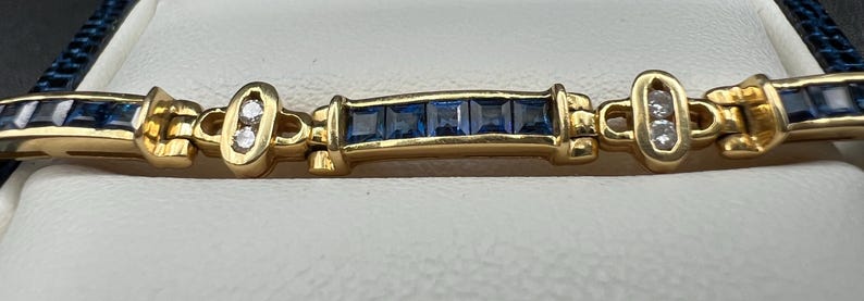 May include: A gold link bracelet featuring alternating sections of channel-set square blue sapphires and oval-shaped gold links, each containing two small round clear diamonds. The bracelet has a polished gold finish and a repeating geometric design.