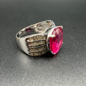 May include: A silver-toned ring featuring a large, oval-cut vibrant pink gemstone as the centerpiece. The band is wide and metallic, accented with multiple rows of small, clear baguette-cut stones set along the sides of the central pink gem.
