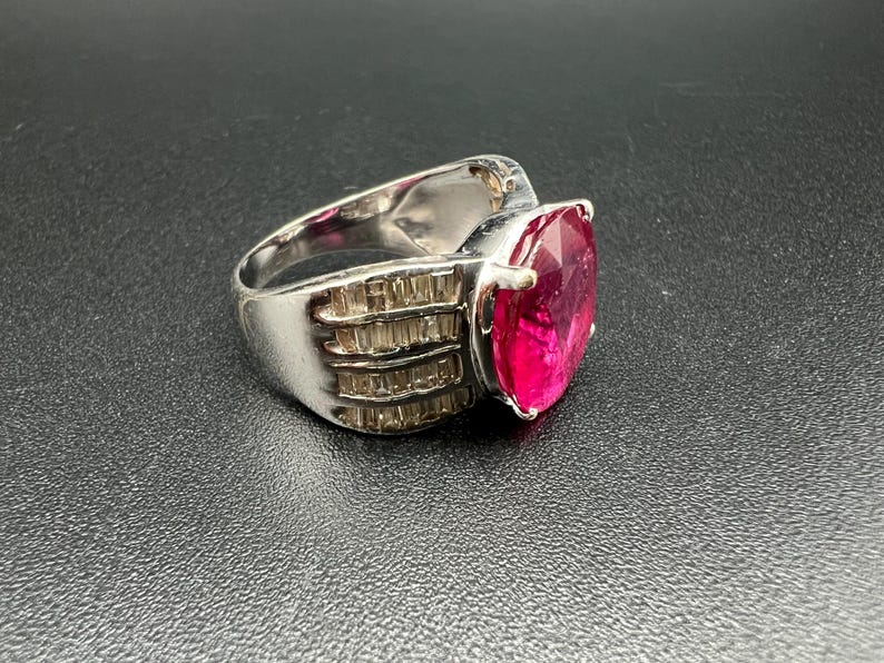 May include: A silver-toned ring featuring a large, oval-cut vibrant pink gemstone as the centerpiece. The band is wide and metallic, accented with multiple rows of small, clear baguette-cut stones set along the sides of the central pink gem.