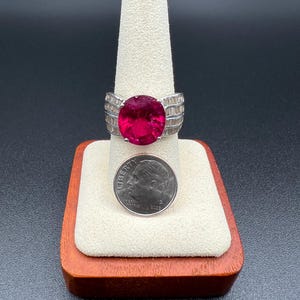 May include: A silver-toned ring featuring a large, oval-cut, deep pink gemstone set in the center, flanked by rows of small, clear baguette-cut stones on the band. A U.S. dime is placed directly below the ring for a clear size comparison.