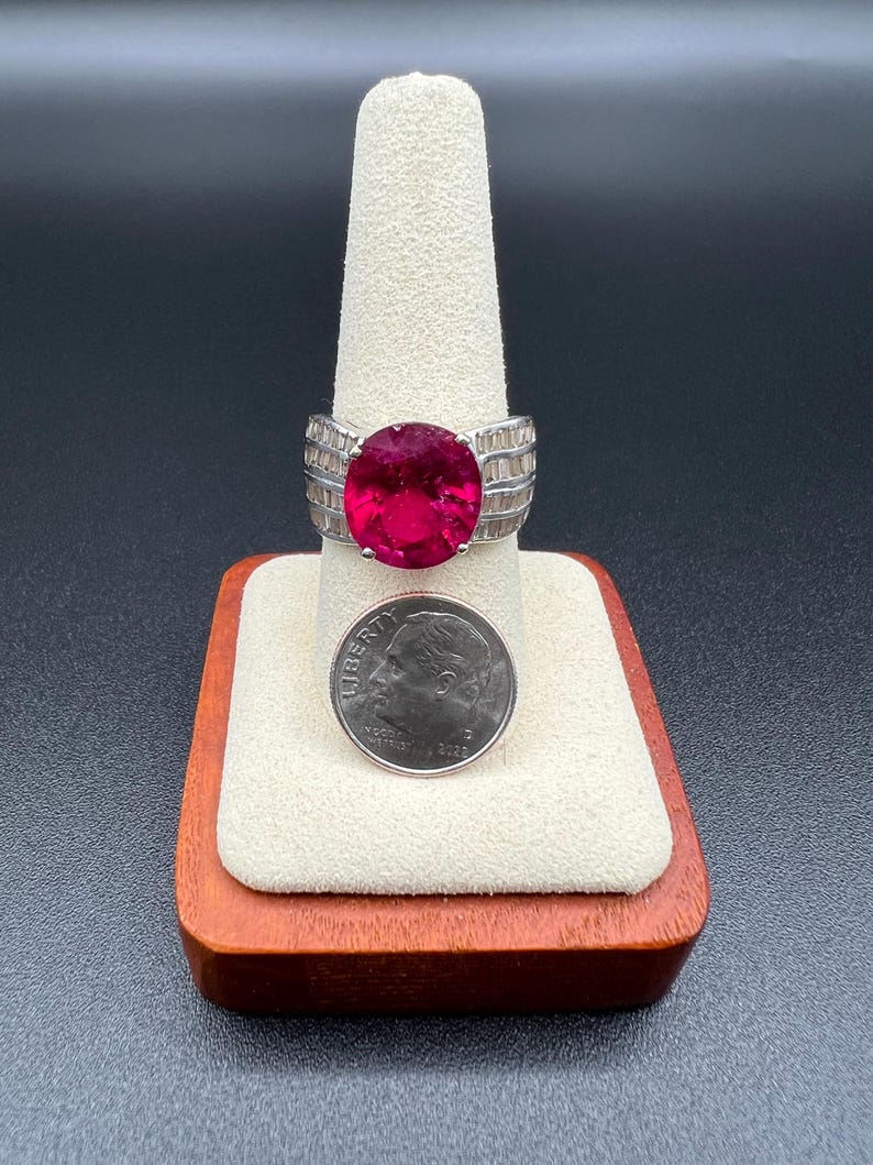May include: A silver-toned ring featuring a large, oval-cut, deep pink gemstone set in the center, flanked by rows of small, clear baguette-cut stones on the band. A U.S. dime is placed directly below the ring for a clear size comparison.
