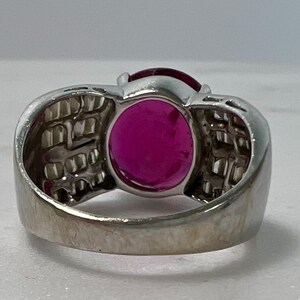 May include: A silver-toned ring featuring a prominent oval-shaped deep pink gemstone at the center. The band is wide and decorated with multiple small, clear rectangular stones set in a grid pattern along the sides of the central setting.