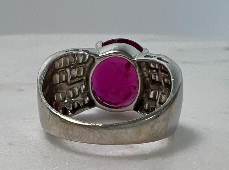 May include: A silver-toned ring featuring a prominent oval-shaped deep pink gemstone at the center. The band is wide and decorated with multiple small, clear rectangular stones set in a grid pattern along the sides of the central setting.