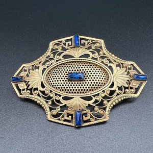May include: Ornate gold-toned brooch with filigree detailing and an oval, perforated center. The brooch features four rectangular, blue gemstones and is set against a dark background. The design includes floral and geometric elements.