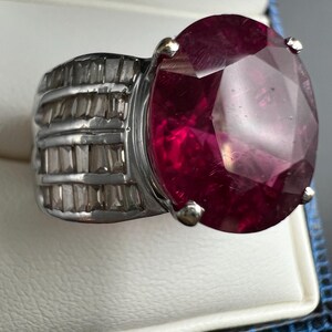 May include: A silver-toned ring featuring a large, oval-cut, deep red gemstone held by four prongs. The band is wide and decorated with multiple rows of small, clear, rectangular baguette-cut stones set horizontally along the sides of the ring.
