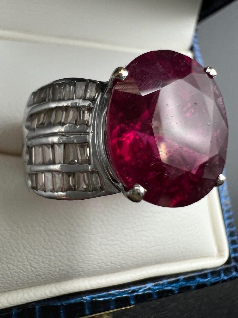 May include: A silver-toned ring featuring a large, oval-cut, deep red gemstone held by four prongs. The band is wide and decorated with multiple rows of small, clear, rectangular baguette-cut stones set horizontally along the sides of the ring.