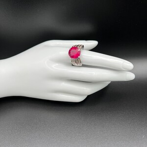 May include: A silver-toned ring featuring a large, round, vibrant red faceted gemstone as the centerpiece. The band is wide and constructed with multiple rows of small, clear, rectangular baguette-cut stones that extend along the sides.