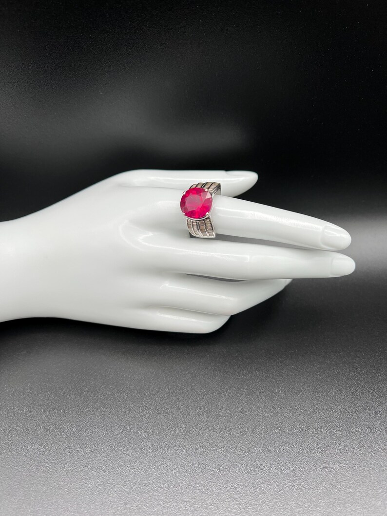 May include: A silver-toned ring featuring a large, round, vibrant red faceted gemstone as the centerpiece. The band is wide and constructed with multiple rows of small, clear, rectangular baguette-cut stones that extend along the sides.