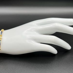 May include: A gold-toned bracelet featuring a series of rectangular blue stones alternating with small, clear, round stones. The design consists of repeating segments of blue and clear stones set in a metallic frame, creating a continuous link pattern.