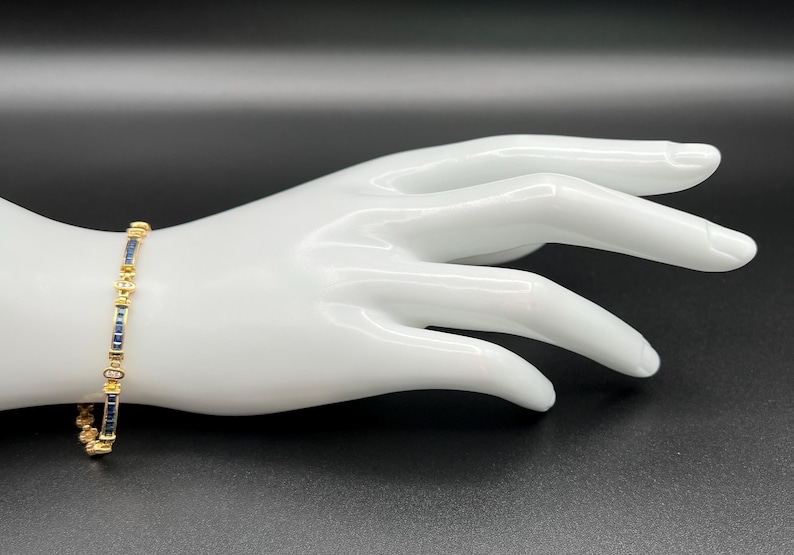 May include: A gold-toned bracelet featuring a series of rectangular blue stones alternating with small, clear, round stones. The design consists of repeating segments of blue and clear stones set in a metallic frame, creating a continuous link pattern.