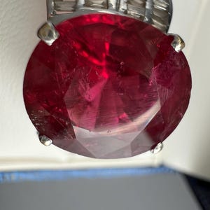 May include: A large, oval-cut, deep red gemstone set in a silver-toned metal ring. The central stone features multiple facets that reflect light, secured by four prominent prongs. The band is accented with a row of smaller, rectangular clear stones.