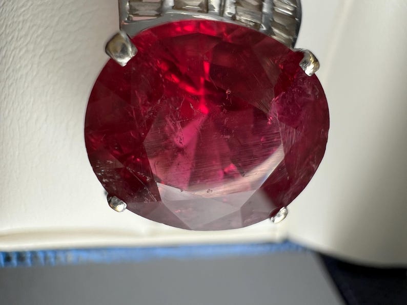 May include: A large, oval-cut, deep red gemstone set in a silver-toned metal ring. The central stone features multiple facets that reflect light, secured by four prominent prongs. The band is accented with a row of smaller, rectangular clear stones.
