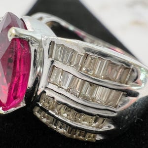 May include: A silver-toned ring featuring a large, faceted, marquise-cut vibrant pink gemstone as the centerpiece. The band is wide and decorated with three parallel rows of small, clear, rectangular baguette-cut stones set in metal channels.