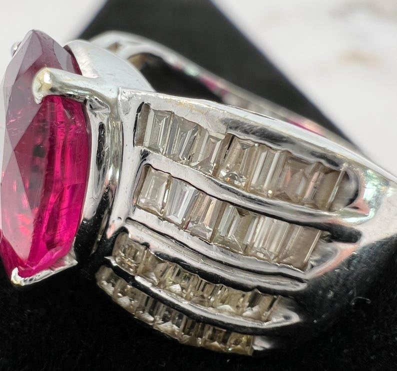 May include: A silver-toned ring featuring a large, faceted, marquise-cut vibrant pink gemstone as the centerpiece. The band is wide and decorated with three parallel rows of small, clear, rectangular baguette-cut stones set in metal channels.