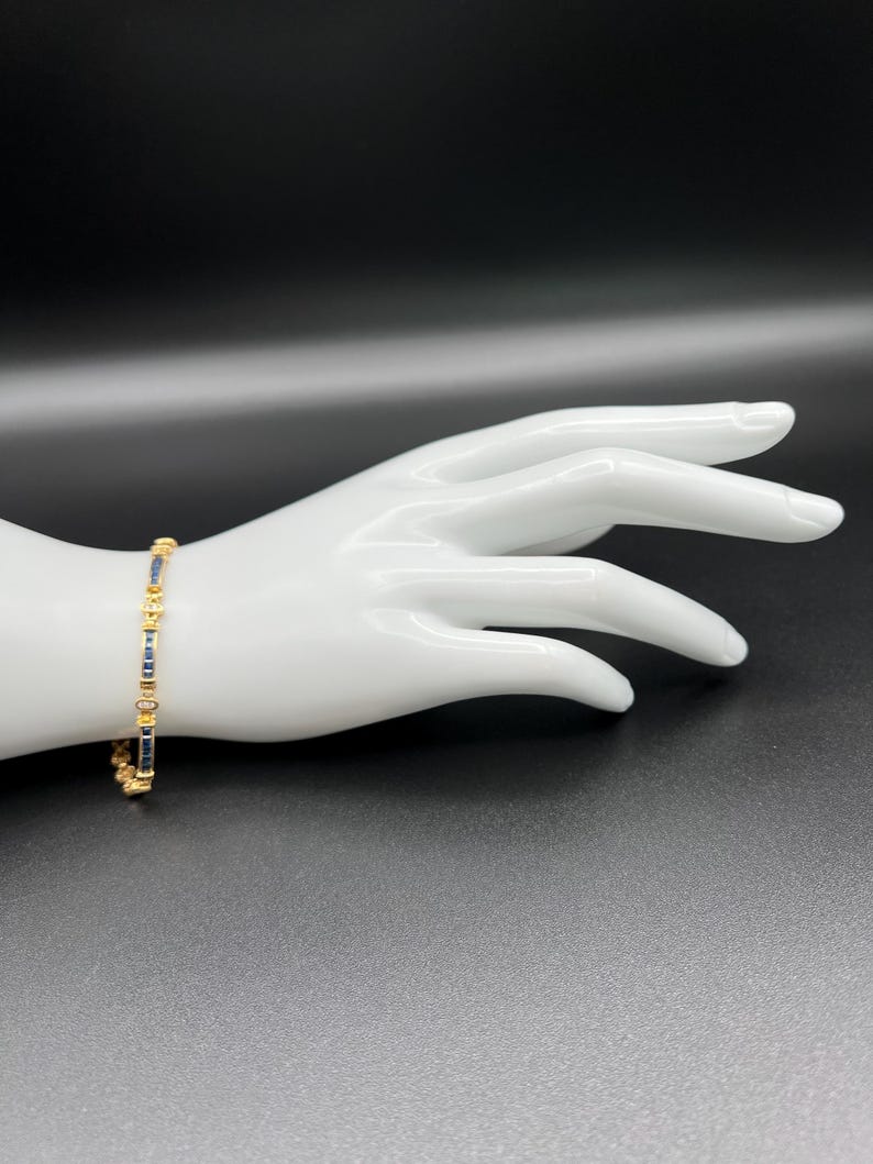 May include: A gold-toned bracelet featuring a repeating pattern of rectangular dark blue stones and small, circular gold-toned accents. The bracelet is constructed with a series of linked segments that alternate between the blue stones and gold metal.