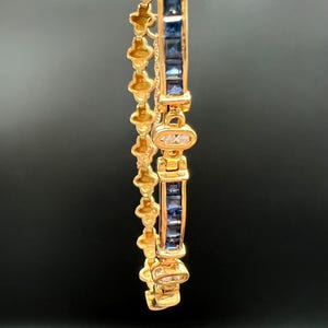 May include: A gold bracelet featuring a series of square-cut blue sapphires set in a channel, alternating with small oval links containing clear stones. The side of the bracelet displays a repeating clover-like pattern in polished yellow gold.