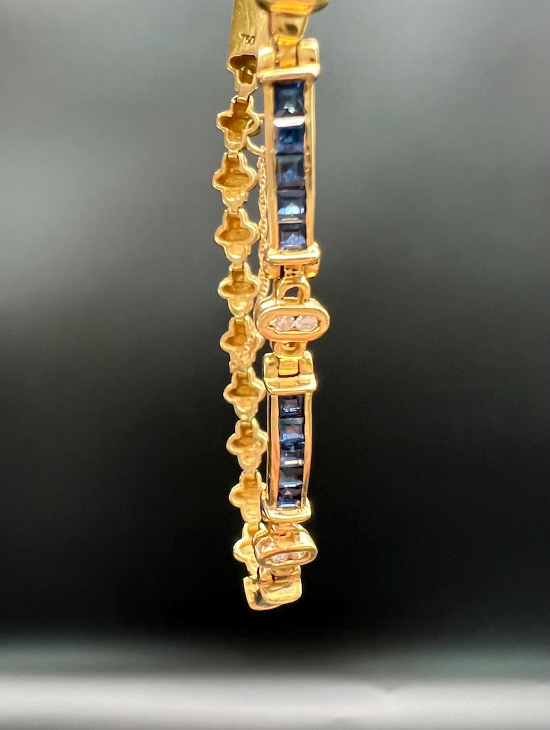 May include: A gold bracelet featuring a series of square-cut blue sapphires set in a channel, alternating with small oval links containing clear stones. The side of the bracelet displays a repeating clover-like pattern in polished yellow gold.