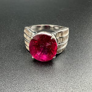 May include: A silver-toned ring featuring a large, round, vibrant pink gemstone set in the center. The band is wide and decorated with multiple rows of small, clear, rectangular baguette-cut stones that extend along the sides of the ring.