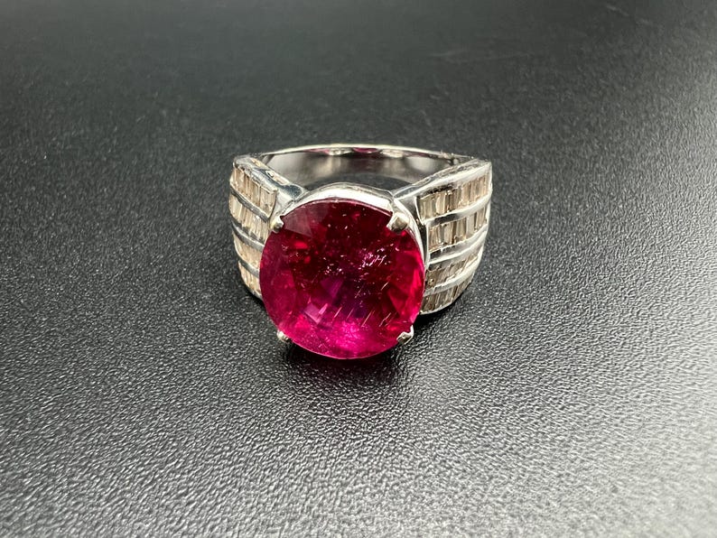 May include: A silver-toned ring featuring a large, round, vibrant pink gemstone set in the center. The band is wide and decorated with multiple rows of small, clear, rectangular baguette-cut stones that extend along the sides of the ring.