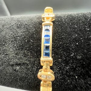 May include: A gold-toned bracelet featuring a central row of four square-cut blue gemstones. The bracelet is constructed with articulated metal links, with small clear stones set into the decorative gold-toned connectors between the blue segments.