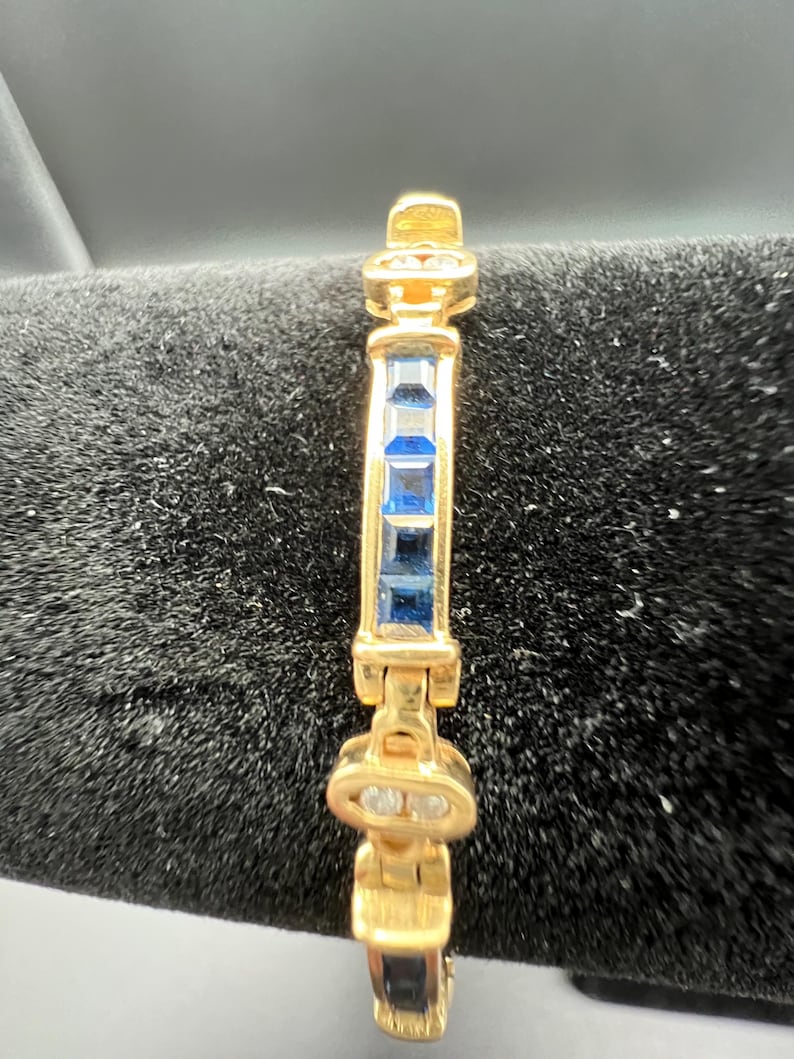 May include: A gold-toned bracelet featuring a central row of four square-cut blue gemstones. The bracelet is constructed with articulated metal links, with small clear stones set into the decorative gold-toned connectors between the blue segments.
