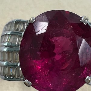May include: A ring featuring a large, round, faceted deep red gemstone set in a silver-toned metal band. The band is accented with multiple rows of small, clear, rectangular baguette-cut stones arranged horizontally along the sides of the setting.