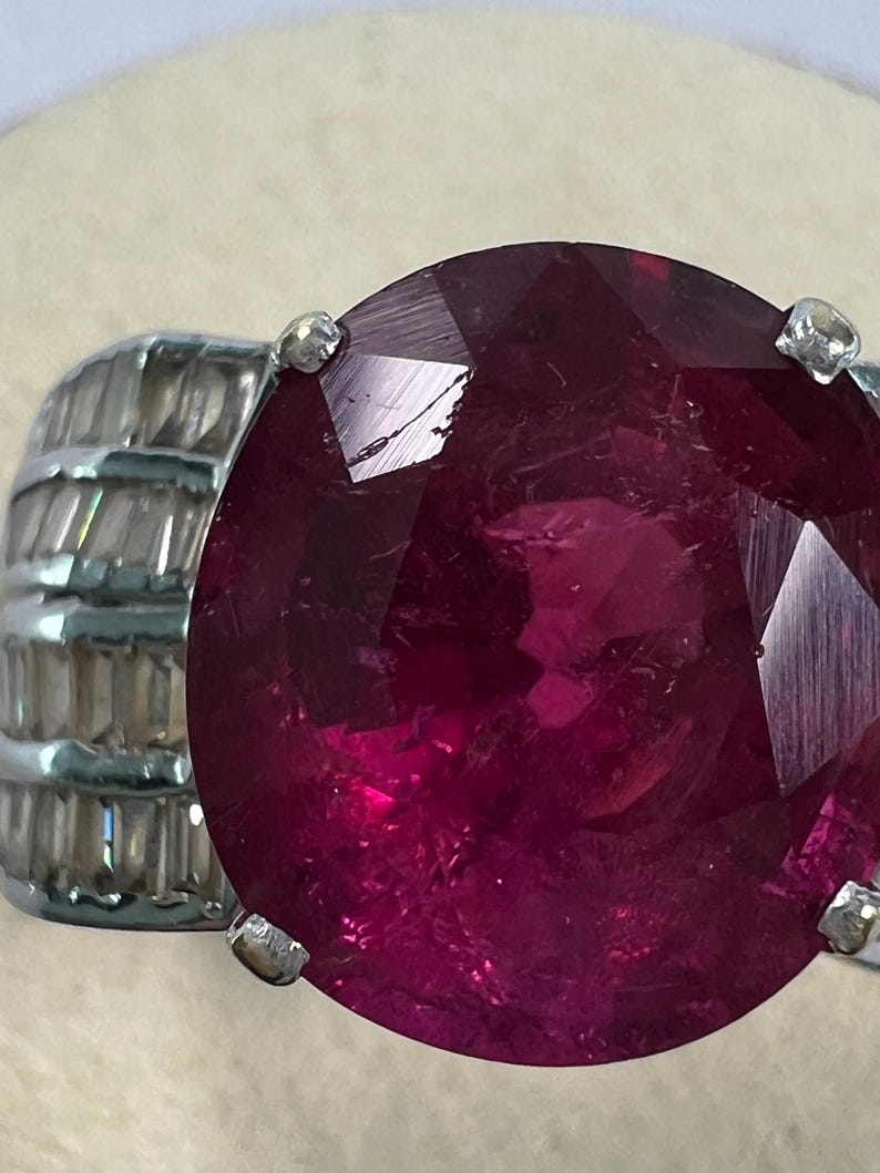 May include: A ring featuring a large, round, faceted deep red gemstone set in a silver-toned metal band. The band is accented with multiple rows of small, clear, rectangular baguette-cut stones arranged horizontally along the sides of the setting.