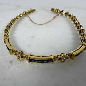 May include: A gold bracelet featuring a series of rectangular links, with alternating sections of channel-set blue sapphires and small clear stones. The piece includes a delicate safety chain and a secure clasp mechanism, creating a continuous circular design.