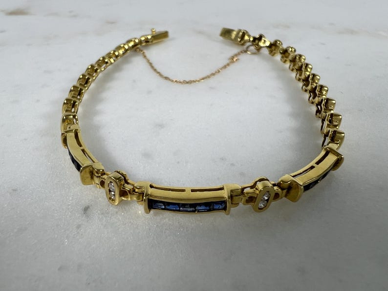 May include: A gold bracelet featuring a series of rectangular links, with alternating sections of channel-set blue sapphires and small clear stones. The piece includes a delicate safety chain and a secure clasp mechanism, creating a continuous circular design.