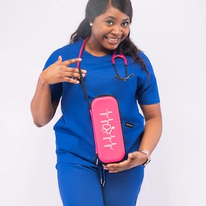 May include: A woman wearing a blue medical scrub top and pants is holding a pink stethoscope case with a white EKG line and stethoscope graphic. The case has the word "NURSE" printed on it.
