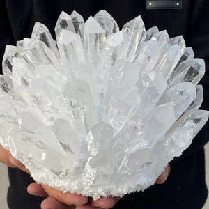 Puede incluir: Un gran grupo de cristales de cuarzo transparente con numerosas formaciones cristalinas puntiagudas. El cuarzo es translúcido, con una base blanca. Los cristales se sostienen en las manos, sobre un fondo oscuro.