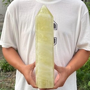 May include: A large, pale yellow crystal tower with a pointed top. The crystal is held by a person wearing a white t-shirt. The crystal's surface has a slightly translucent appearance.