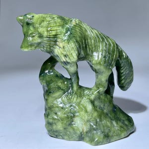 Puede incluir: Una escultura de lobo de jade verde de pie sobre una roca. El lobo está tallado en un estilo realista y tiene una cara detallada. La roca también está tallada con detalle y tiene una textura rugosa.