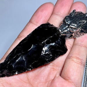 Puede incluir: Un colgante de punta de flecha de obsidiana negra con un dije de cabeza de lobo plateado unido a una cadena plateada.