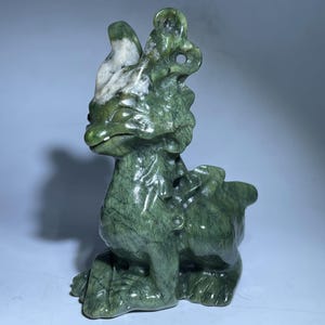May include: A green jade dragon figurine with white markings. The dragon is sitting on its hind legs with its front paws tucked in. It has a long tail and a small, pointed head.