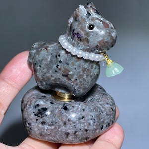 May include: A grey and speckled stone sculpture of a horse, adorned with a pearl necklace and a green tassel pendant. The horse sits atop a round base with a gold-coloured metal accent.