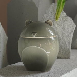 Può includere: Un vaso di ceramica grigia con coperchio, con un design a forma di orso con piccole orecchie. Il vaso è posto su uno sfondo di blocchi di pietra grigia e tulipani rosa, creando un'estetica serena e naturale.