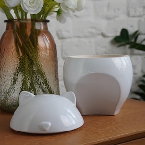 Può includere: Una natura morta con un bouquet di fiori bianchi in un vaso di vetro ambrato strutturato. Un vaso di ceramica bianca e un coperchio abbinato a forma di testa di gatto sono su una superficie di legno. Lo sfondo è un muro di mattoni bianchi.