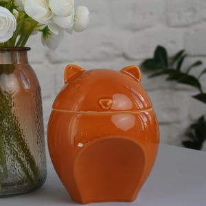 May include: A clear glass vase with a textured surface holds white flowers. Next to it is an orange ceramic jar with a lid, shaped like a bear. The background features a white brick wall and greenery.