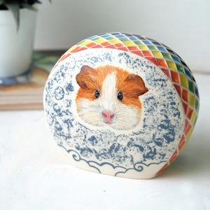 以下が含まれることがあります： 手描きの顔が特徴的な、セラミック製のモルモットの置物。体は白地に青い花柄、上部はカラフルなモザイク模様で装飾されています。ユニークな装飾品です。