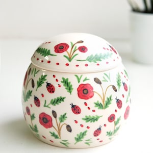 Puede incluir: Un tarro de cerámica redondo con tapa a juego, que presenta una base de color crema decorada con amapolas rojas pintadas a mano, follaje verde y pequeñas mariquitas rojas esparcidas por toda la superficie del cuerpo y la tapa.