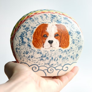 Puede incluir: Un recipiente de cerámica redondo pintado a mano con un diseño de Cavalier King Charles Spaniel. El recipiente tiene un fondo moteado azul y blanco, un borde arcoíris y un diseño decorativo en forma de nube en la base.
