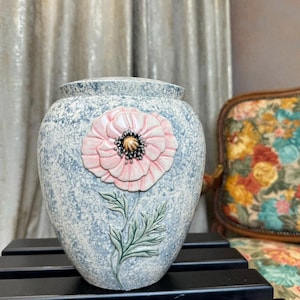 May include: A ceramic vase with a blue and white speckled glaze. The vase features a raised pink poppy flower with a yellow center.